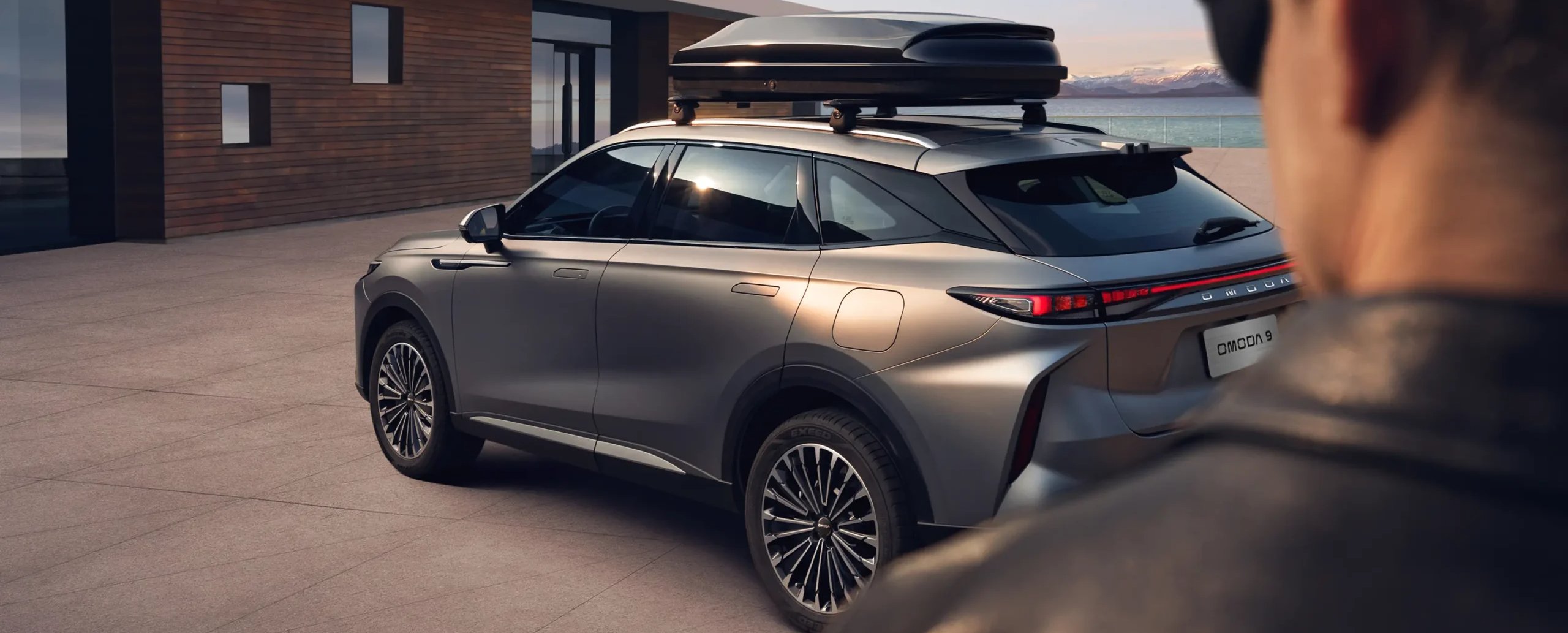 Silver SUV with a roof box parked near a modern house, person in foreground.