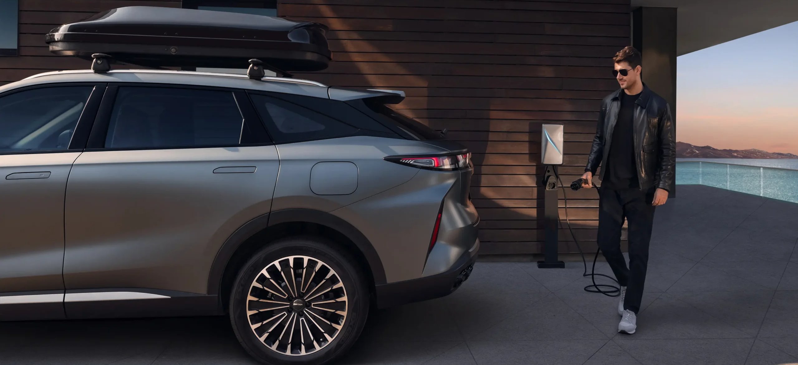 Man in sunglasses walking towards a grey SUV with a rooftop box, holding an electric charging plug.
